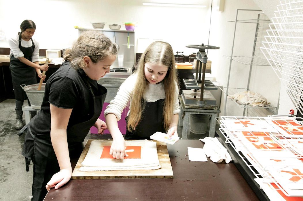 Zwei Mädchen beugen sich über einen Tisch in der Druckwerkstatt, wo sie einen Druck des Logos des Museums herstellen, im Hintergrund rührt ein weiteres Mädchen in einem Zuber
