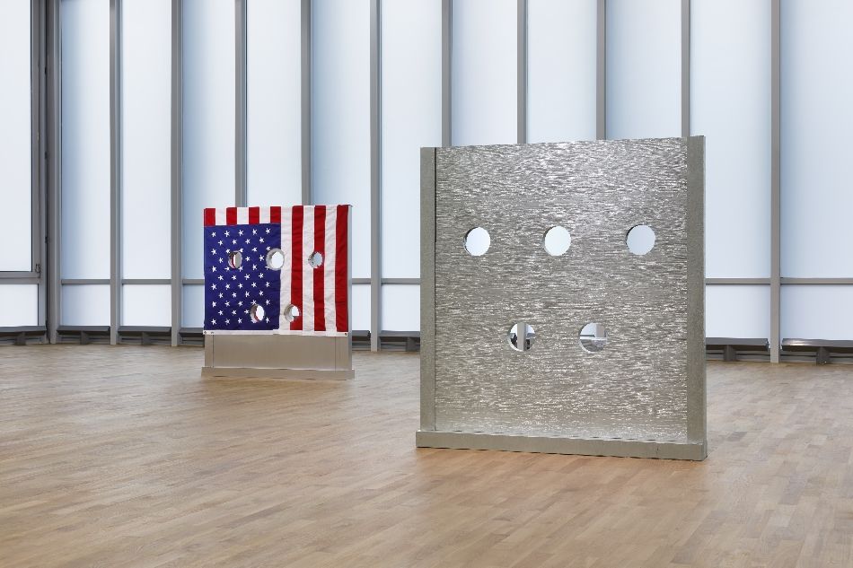 Zwei Kunstwerke von Cady Noland, die Pranger nachbilden und abwandeln, ein silberner im Vordergrund rechts, einer mit der amerikanischen Flagge bedruckter im Hintergrund links