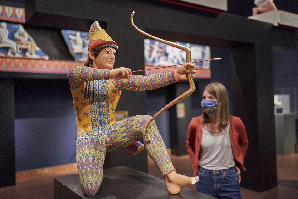Frau mit Maske betrachtet bunte Skulptur eines knieenden Kriegers mit Pfeil und Bogen in der Ausstellung im Liebieghaus