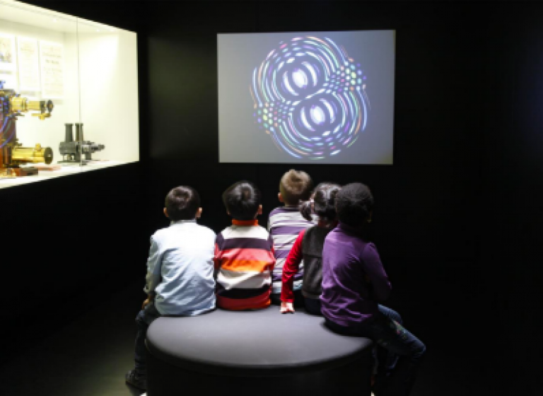 Kinder sitzen vor Bildschirm und Vitrine in einem abgedunkelten Raum im Deutschen Filmmuseum